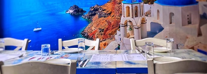 A laid table at Zorba The Greek in front of the Santorini mural — blue sea, caldera cliffs and whitewashed buildings overlooking the water
